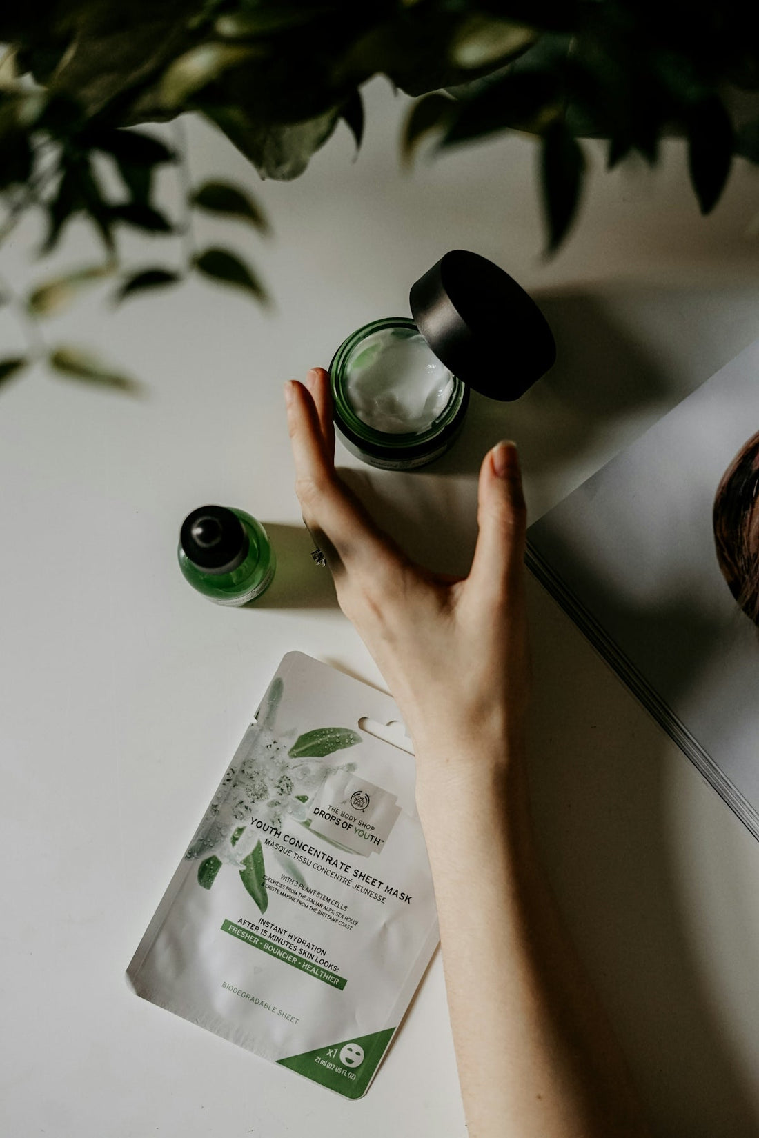 A hand reaching toward a jar of cream, a serum bottle, and a sheet mask